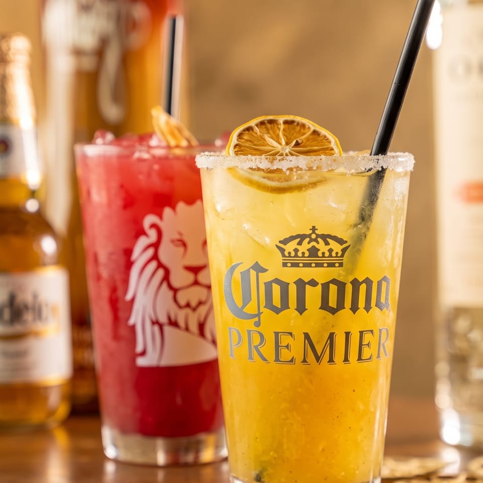 Citrus cocktail with a sugar rim, dried lemon garnish, and straw beside a red drink on a warmly lit bar.