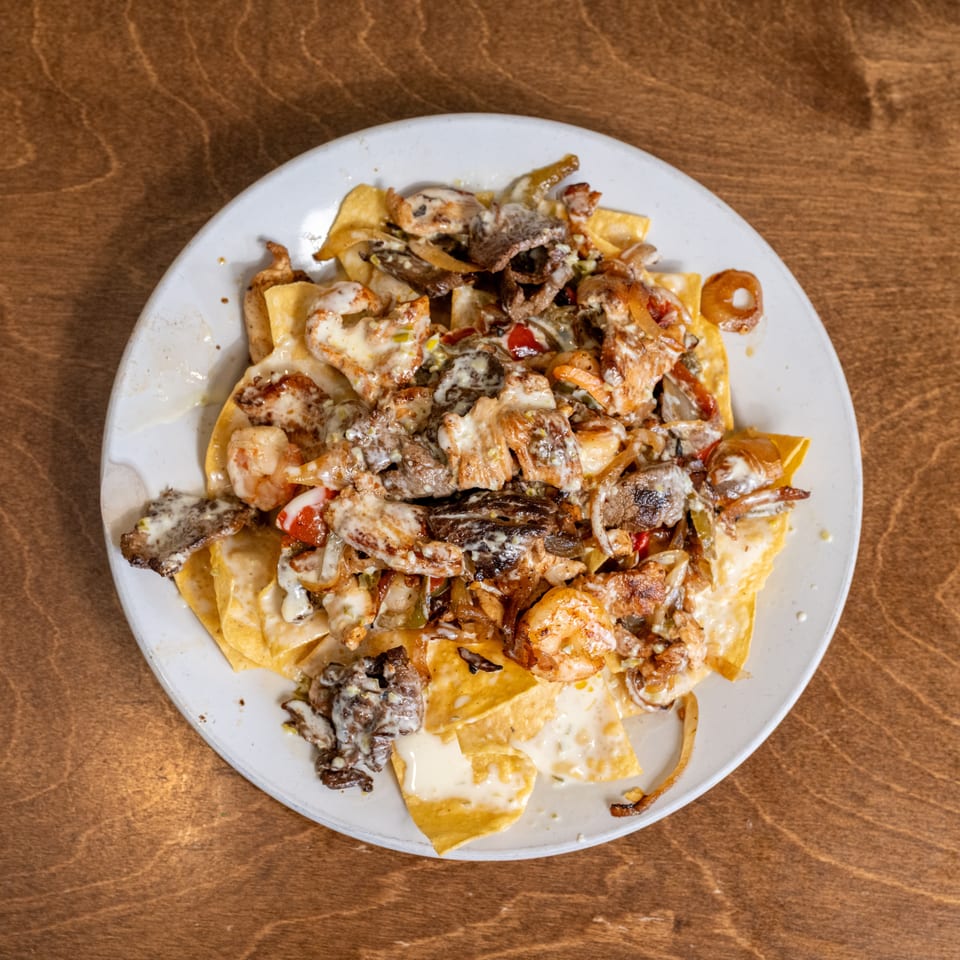Loaded nachos topped with shrimp, grilled meats, vegetables, and creamy sauce on a white plate.