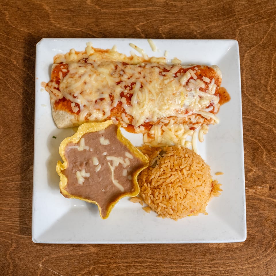 Cheesy enchiladas served with Mexican rice and refried beans on a white plate atop a wooden table.