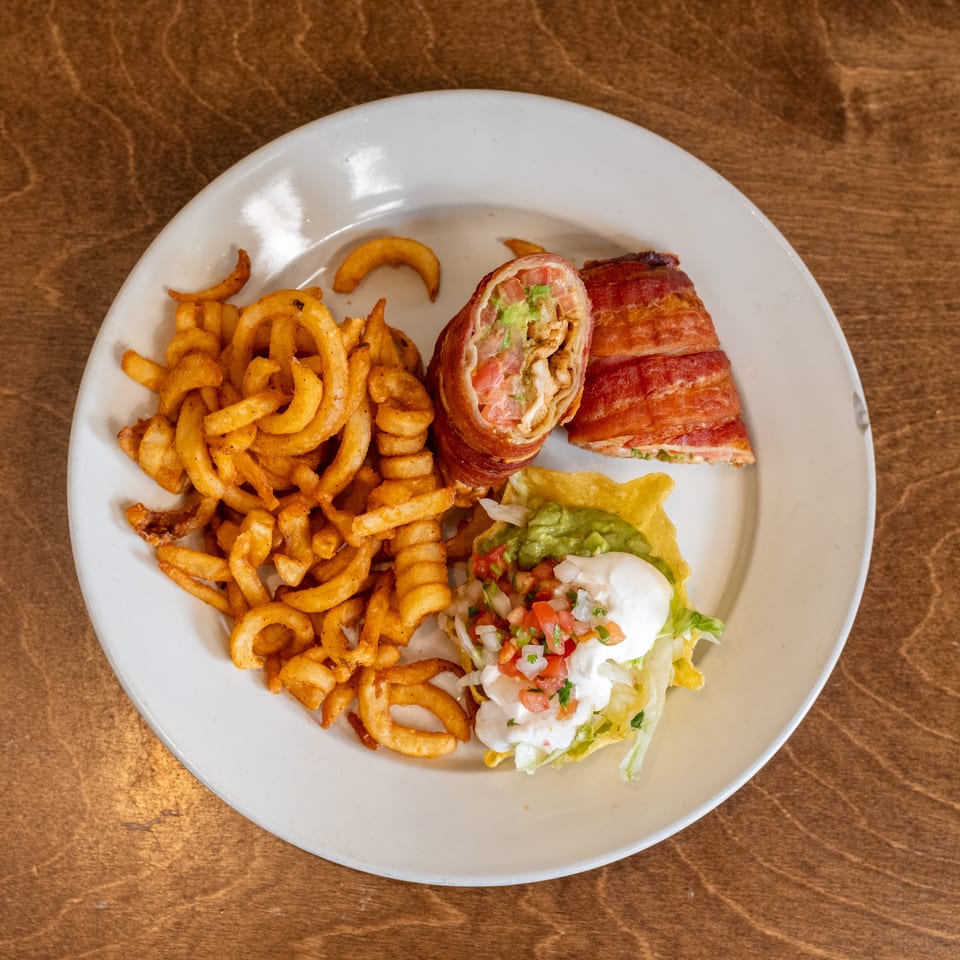 Plate of bacon-wrapped stuffed wraps, curly fries, and a lettuce taco topped with guacamole, salsa, and sour cream.
