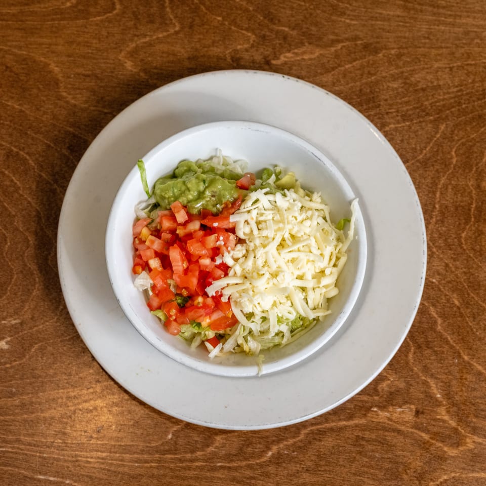 Overhead view of a Mexican-style salad topped with guacamole, diced tomatoes, shredded cheese, and lettuce on a wooden table.