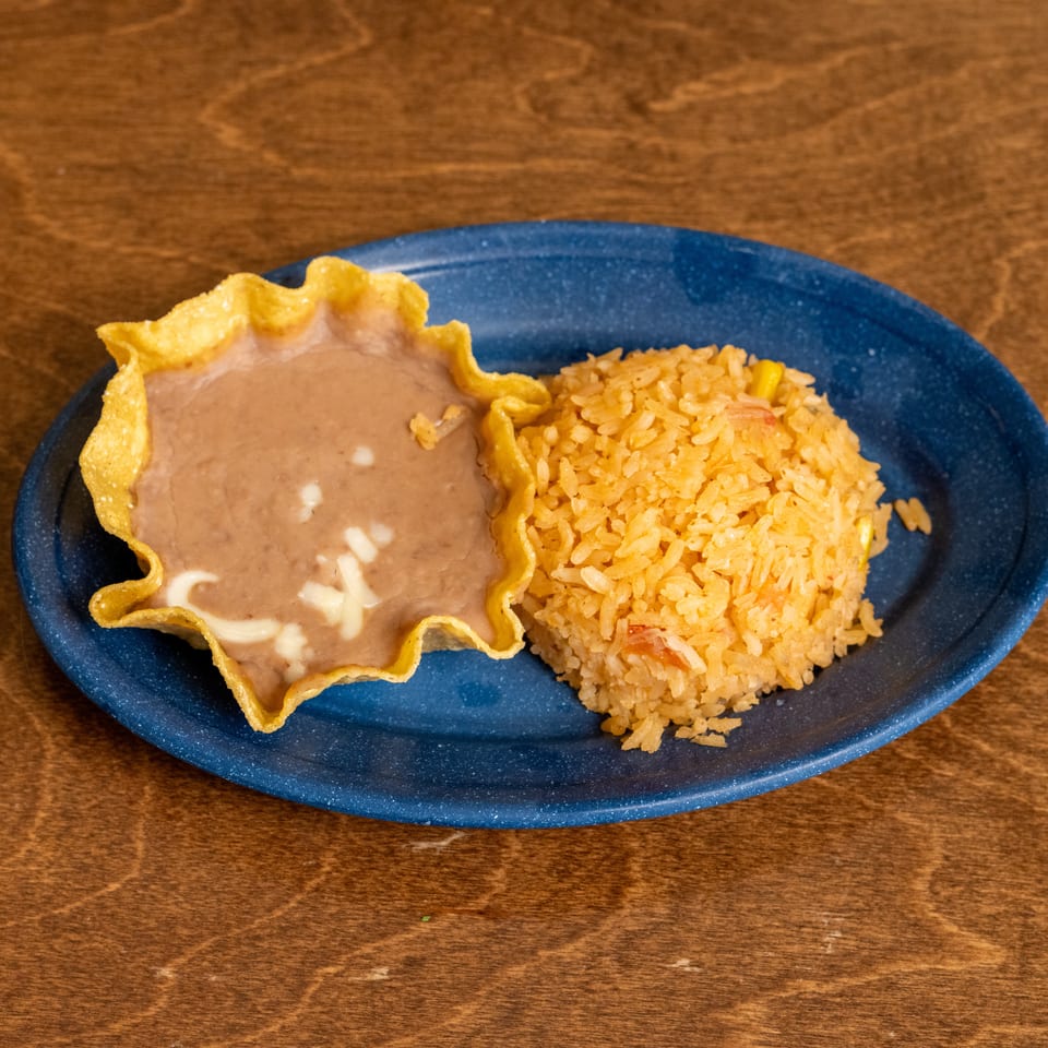 Blue plate holding a crispy tortilla bowl of refried beans and a mound of orange Mexican rice on a wooden table.