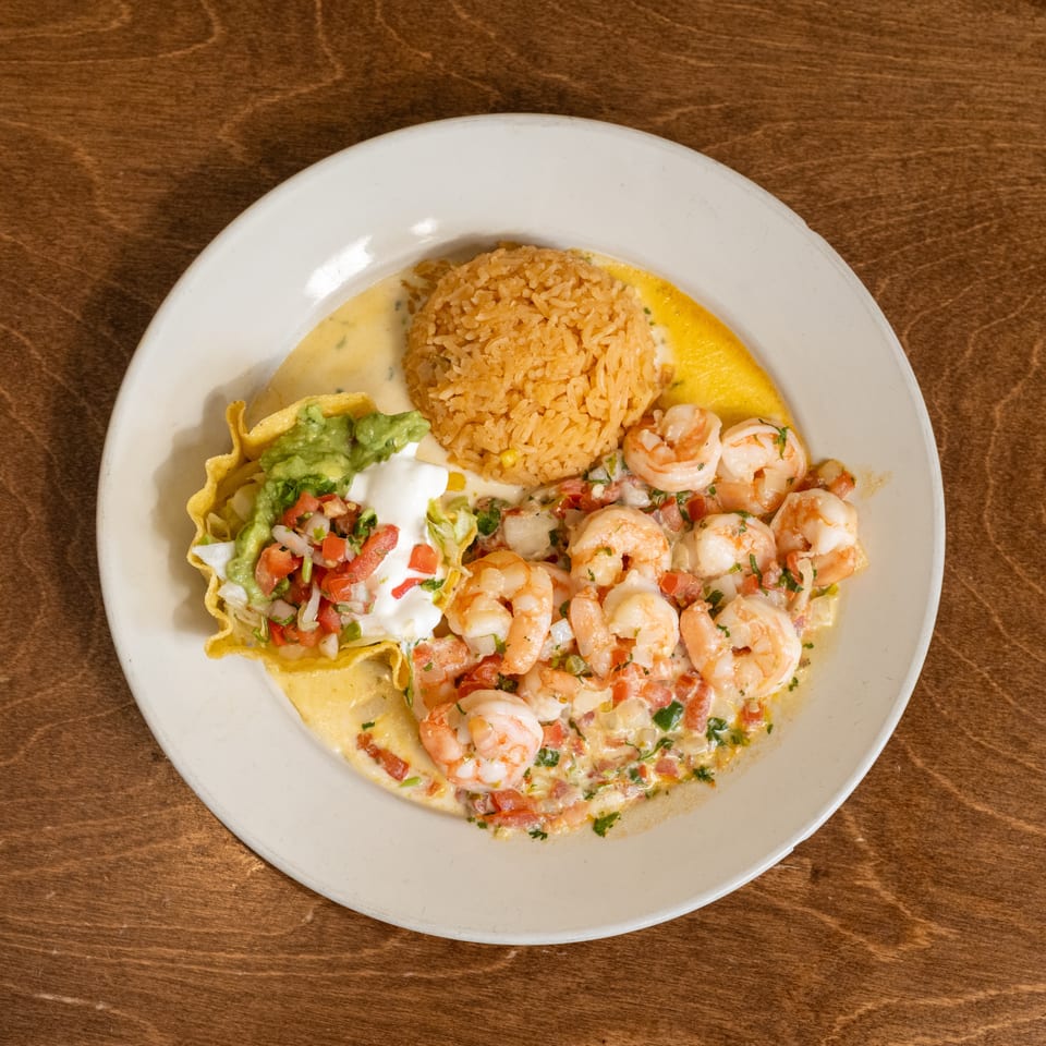 Plate of shrimp in creamy sauce with Mexican rice, guacamole, pico de gallo, and tortilla chips on a wooden table.