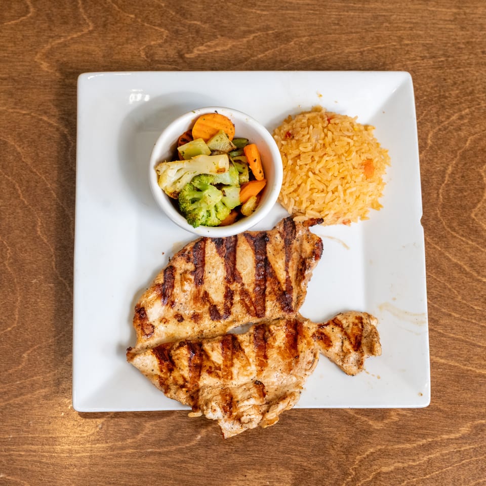 Grilled chicken breasts served with seasoned rice and a bowl of roasted mixed vegetables on a white plate.